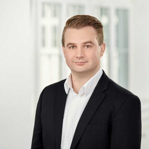 Mann mit kurzen, blonden Haaren, trägt einen schwarzen Anzug und ein weißes Hemd, steht vor einem hellen Hintergrund.