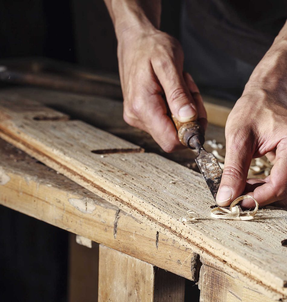 Hände eines Handwerkers, die mit einem Schnitzwerkzeug an einem Holzbrett arbeiten.